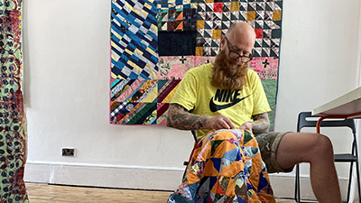 A man sat on a stool stitching a quilt
