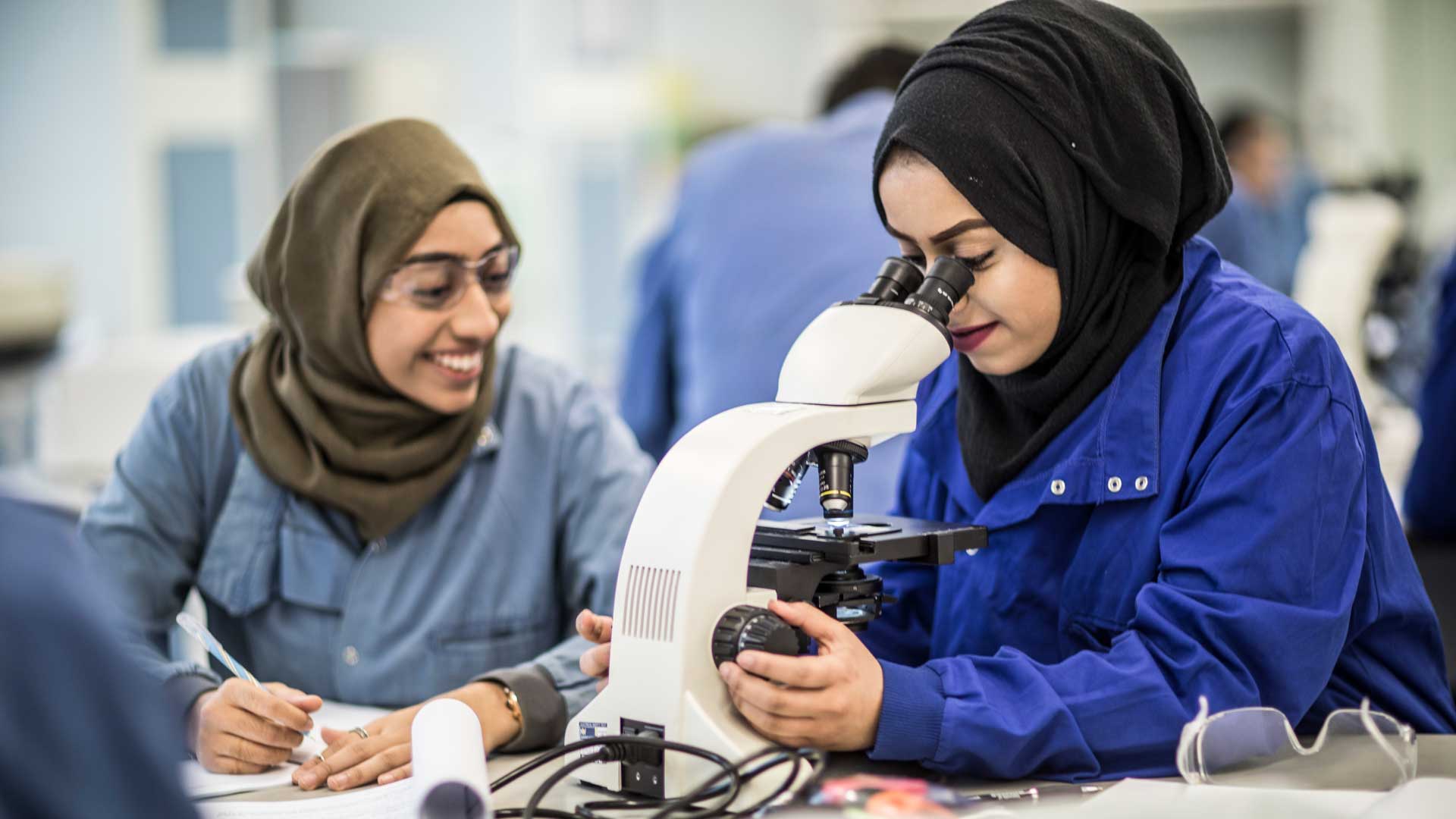 Two students in a biology class. One looking into a microscope while the other takes notes. 