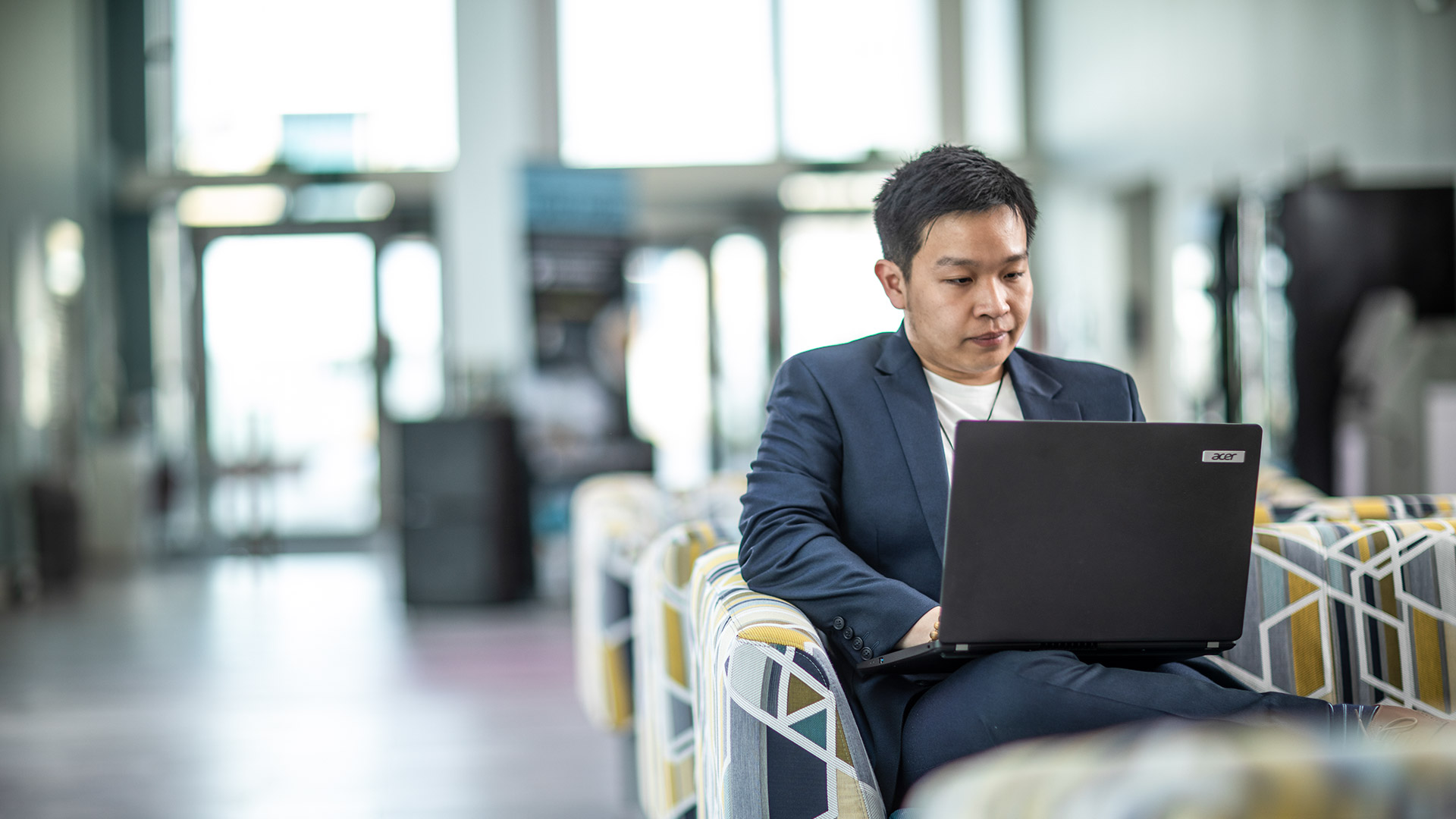 A student wearing a blazer, working on a laptop in a large open communal space .