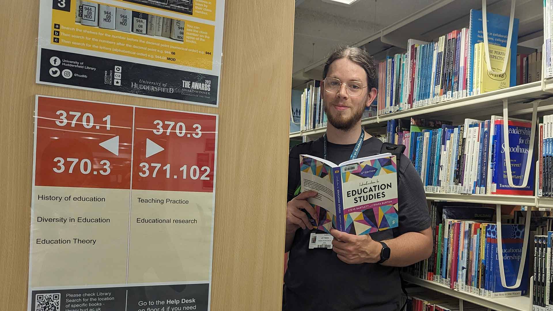 Joe stood in a library aisle, holding an education textbook.